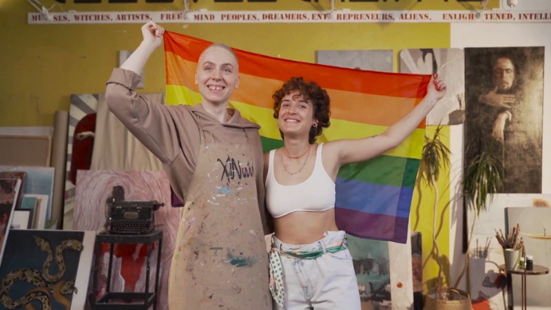 The image shows two people holding a rainbow flag, smiling in an art studio