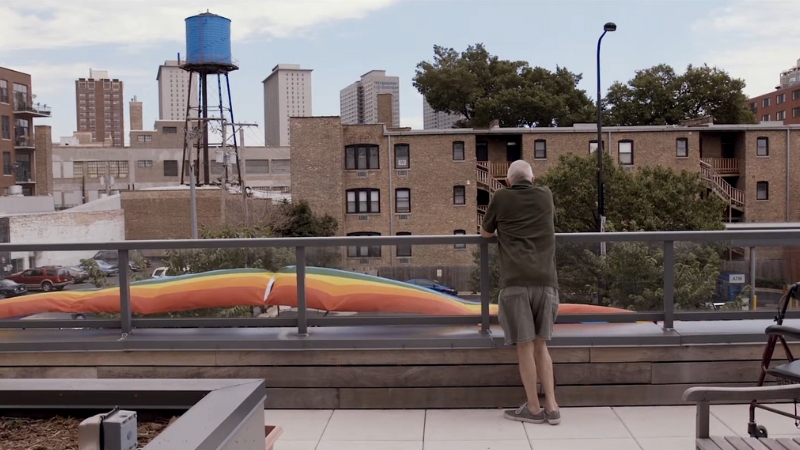 A person stands on a balcony overlooking the city, next to a rainbow banner