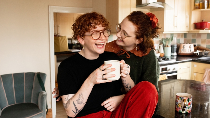 Two queer roommates share a laugh in their kitchen
