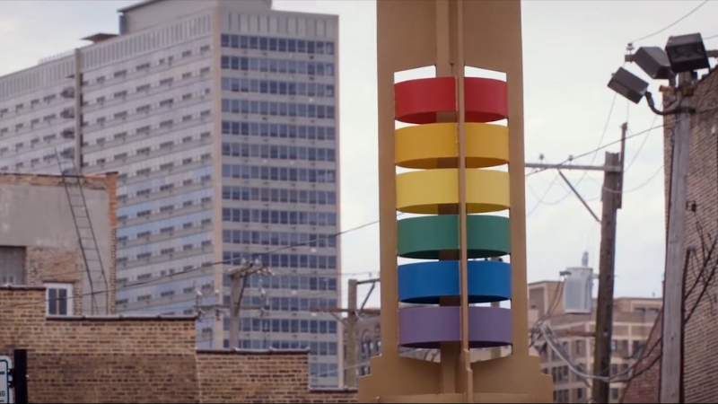 A rainbow-colored public art piece stands in front of city buildings, symbolizing support for queer-friendly housing in Tucson
