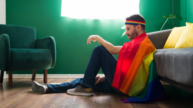 A man sits on the floor wrapped in a pride flag, reflecting concern and caution when searching for queer-friendly housing
