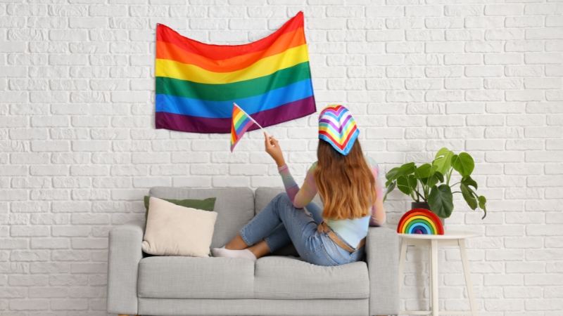A person sits on a couch holding a pride flag, with a rainbow flag on the wall