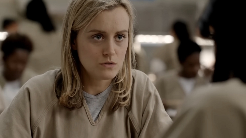A woman in a beige prison uniform sits at a table, portraying a scene from the Netflix series Orange Is the New Black