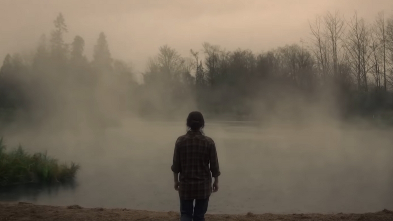 A person stands alone by a fog-covered lake in a haunting scene from the Netflix series The Haunting of Bly Manor