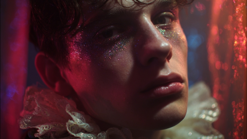 A person with glitter on their face looks into the light, wearing lace around their neck