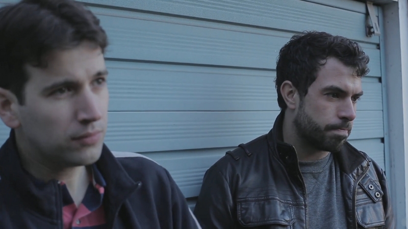 Two men stand outside near a blue wall, sharing a quiet moment in a scene from Weekend (2011)