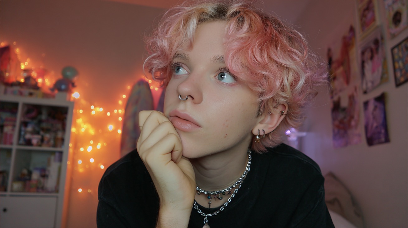 Person with short curly pink hair resting their chin on their hand in a softly lit bedroom with string lights in the background