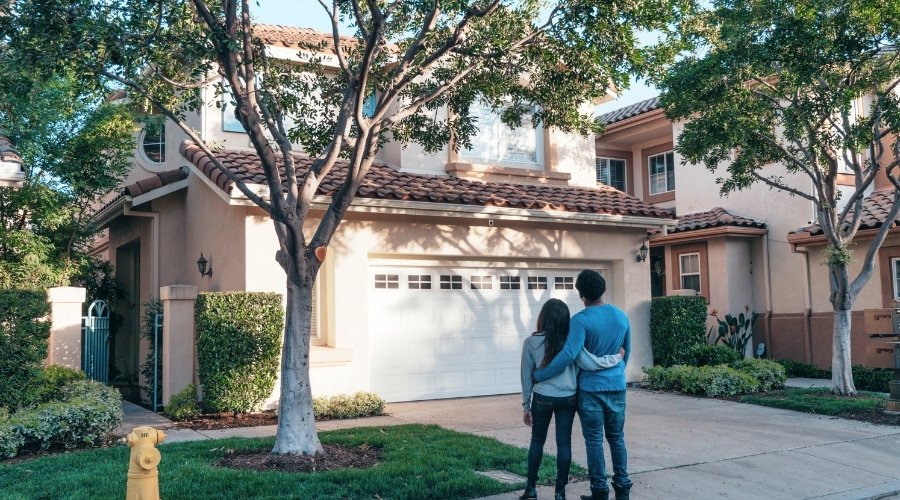 Couple standing together in front of a suburban home, considering buying a house together.